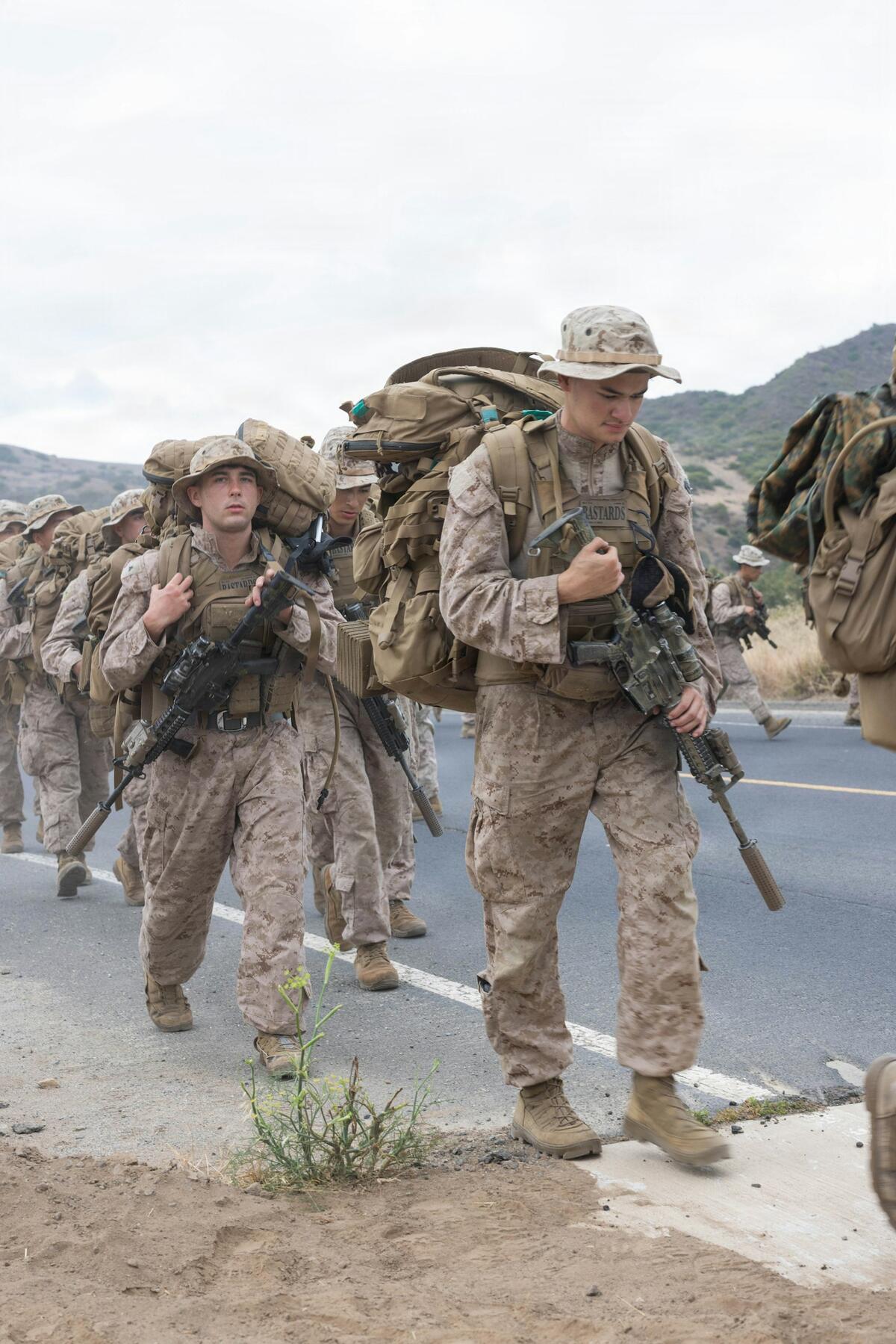 Soldiers in camouflage with heavy packs and rifles march along a road