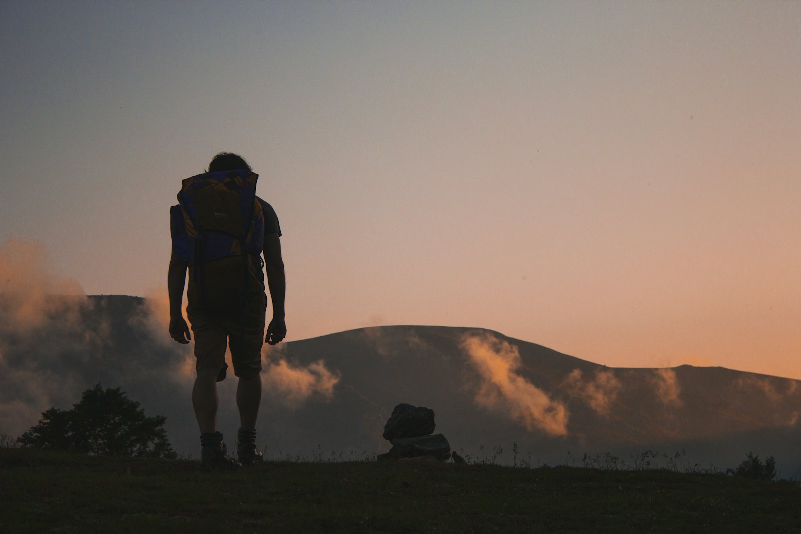 Silhouette of a man with a loaded pack, seen from behind, on a grassy rise in front of a hill at dusk