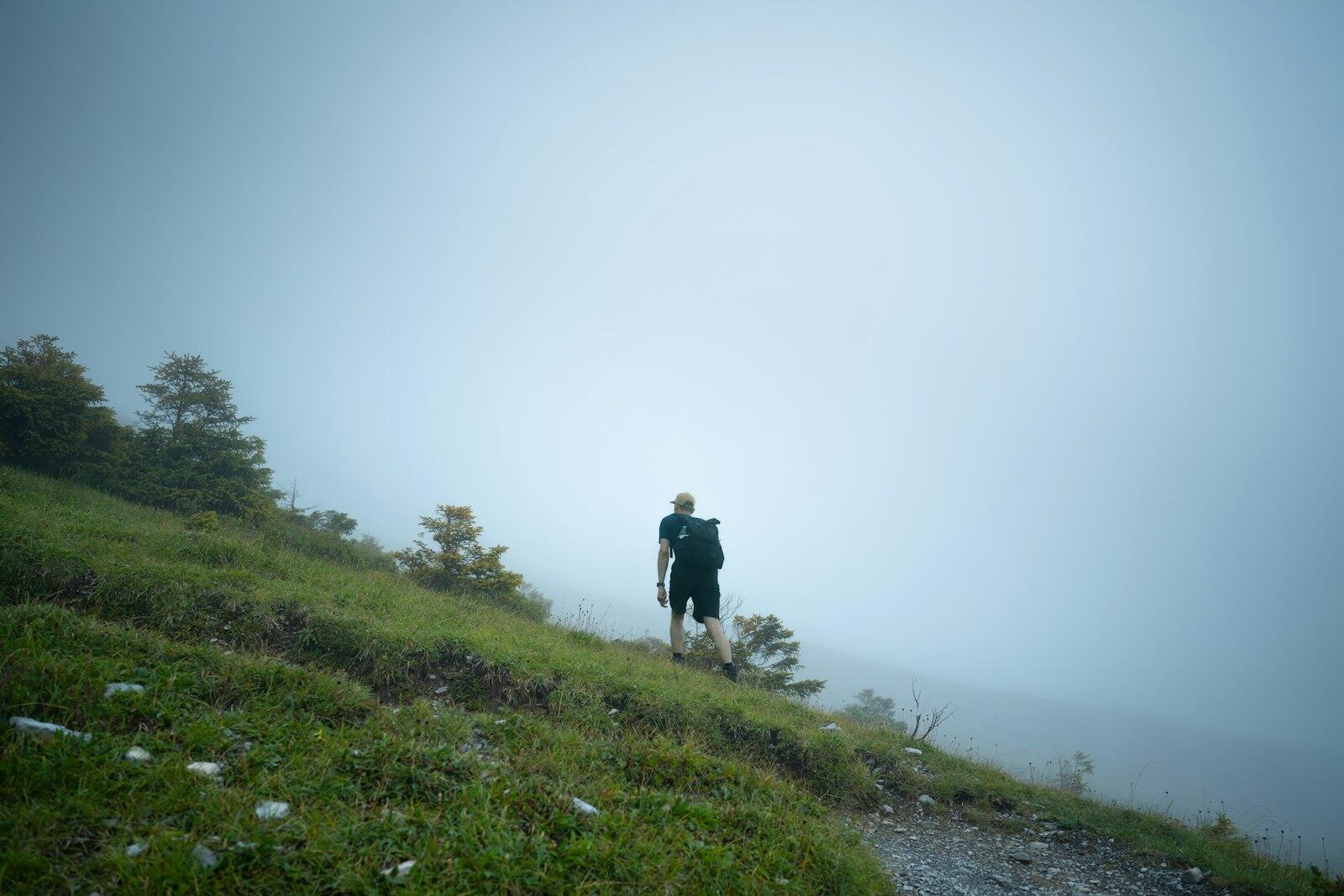 A hiker with a backpack follows a path along a foggy grass slope