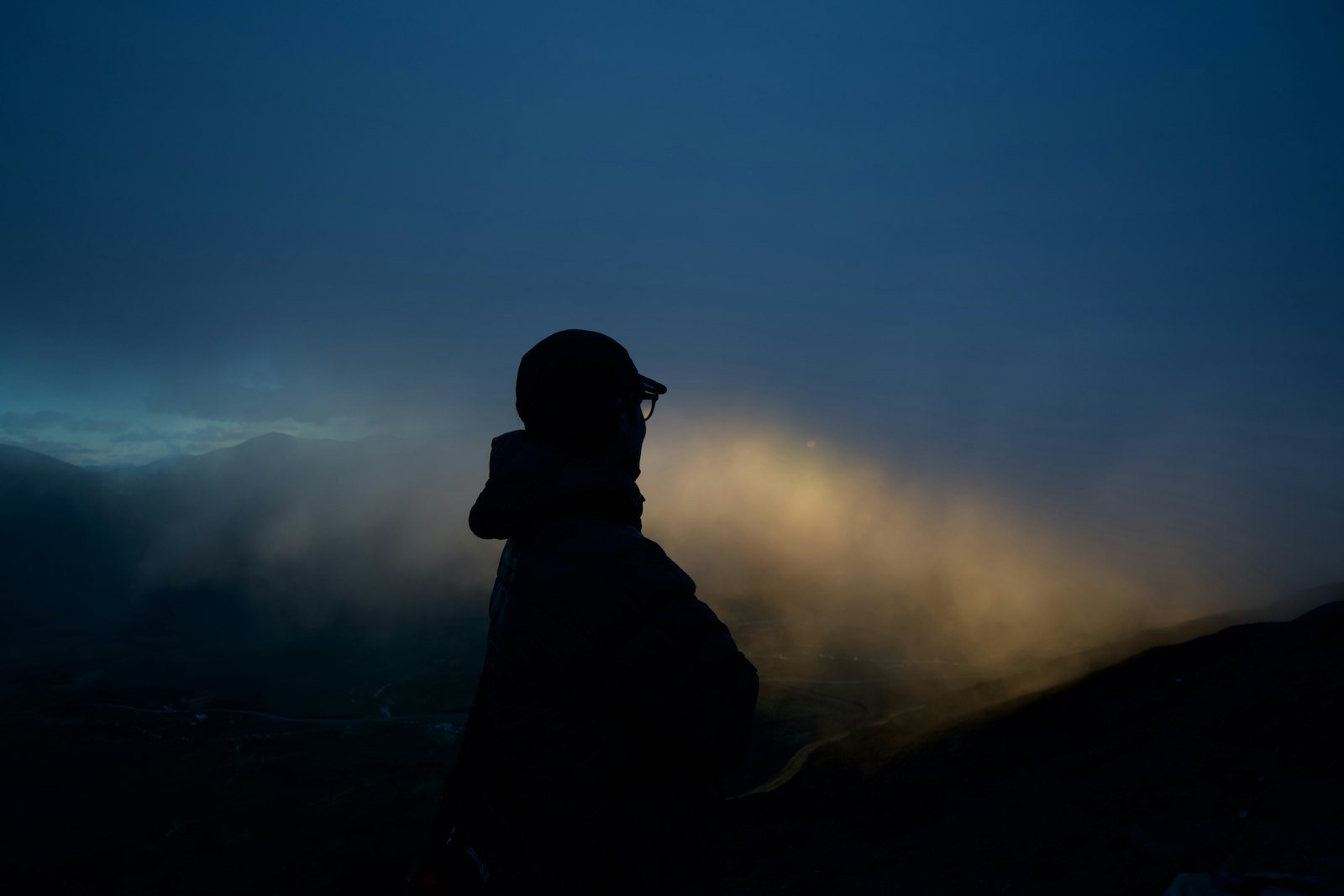 Dark silhouette of a person in a cap on a hillside at dusk, a light beam through the mist behind them