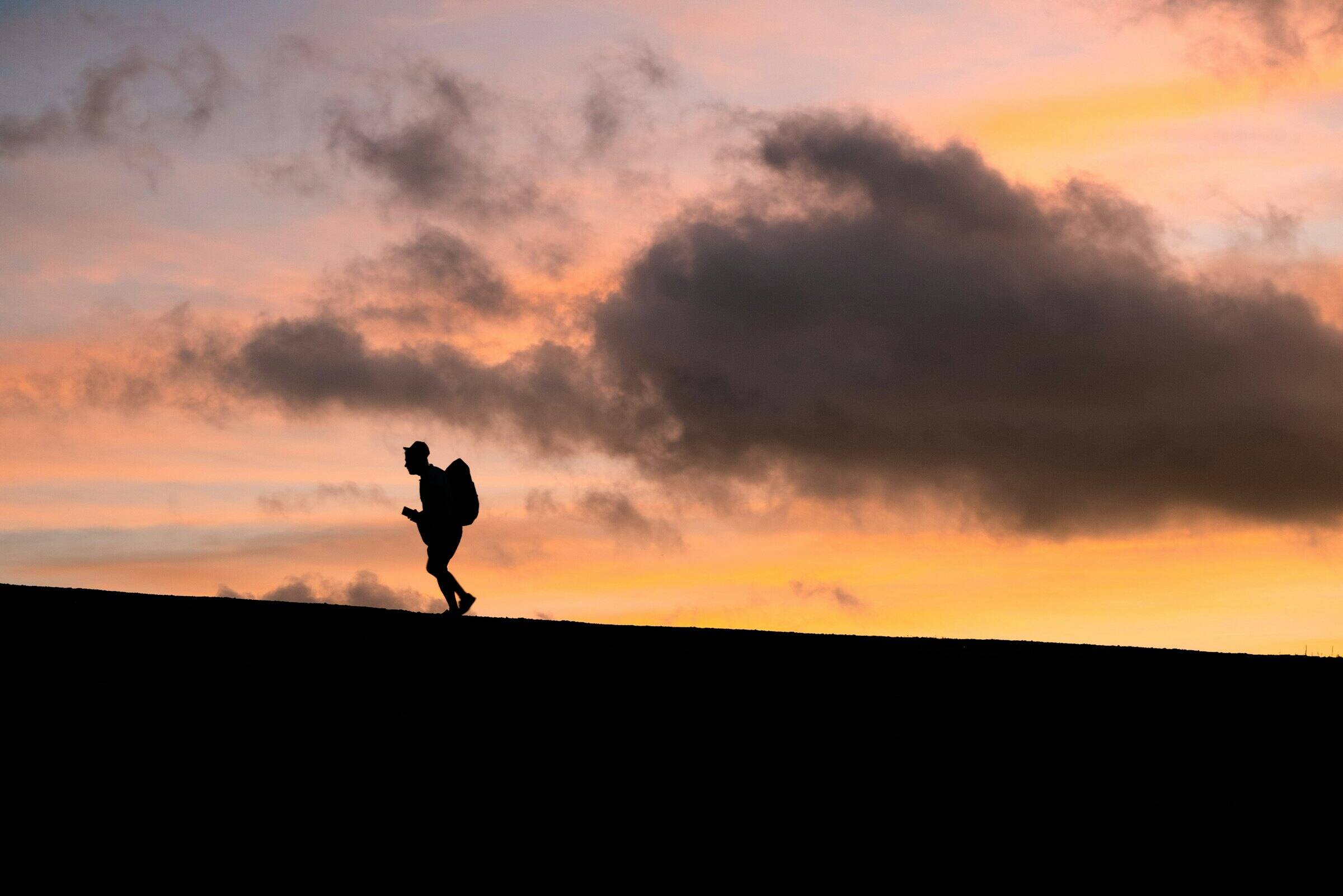 Silhouette of a hiker with a pack walking up a hill against an orange dusk sky