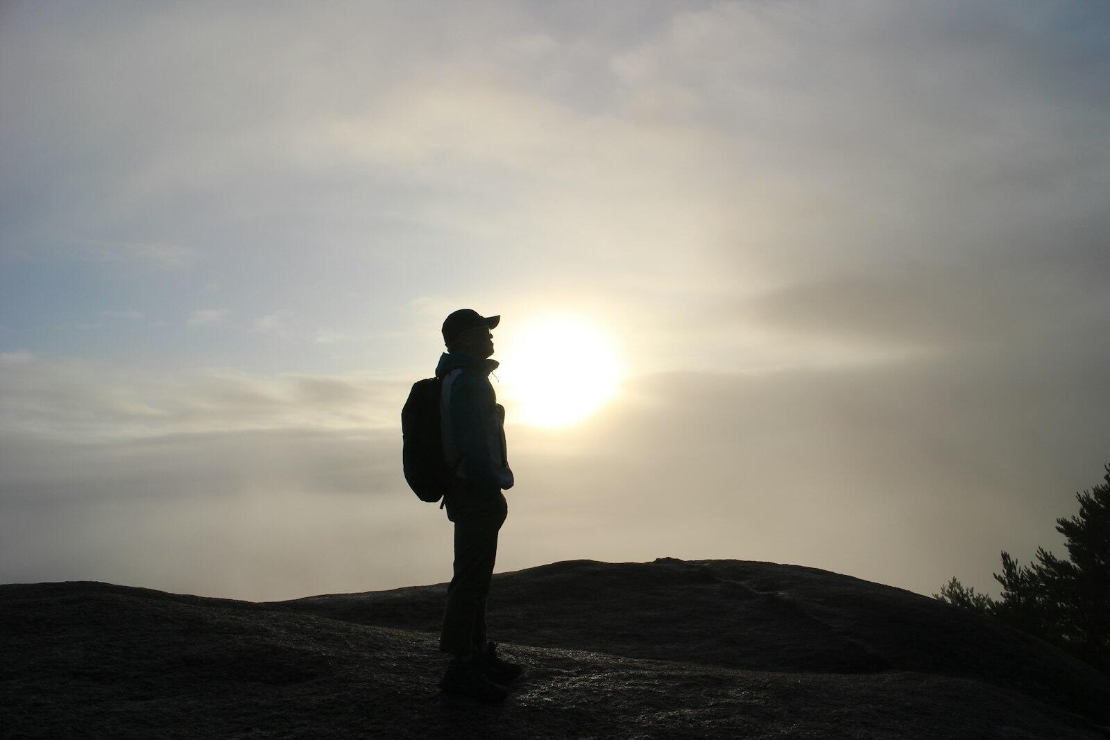 Silhouette of a person with a backpack standing on a ridge in front of the low sun